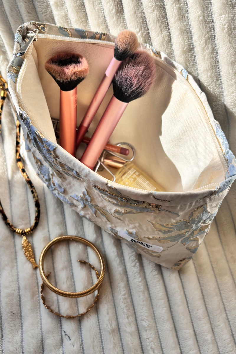 Fency Toilettas van Fencyjewels met gouden bloemenprint, gevuld met make-upkwasten en sieraden op een zacht gestreept kleed. Luxe toilettasje voor sieraden en beauty essentials.