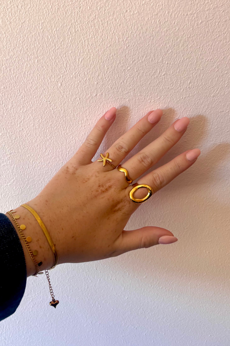 Hand met gouden Fencyjewels ringen, waaronder de Beachy Ring met zeester en schelp design, gecombineerd met minimalistische armbanden — zomerse sieraden look van Fencyjewels.