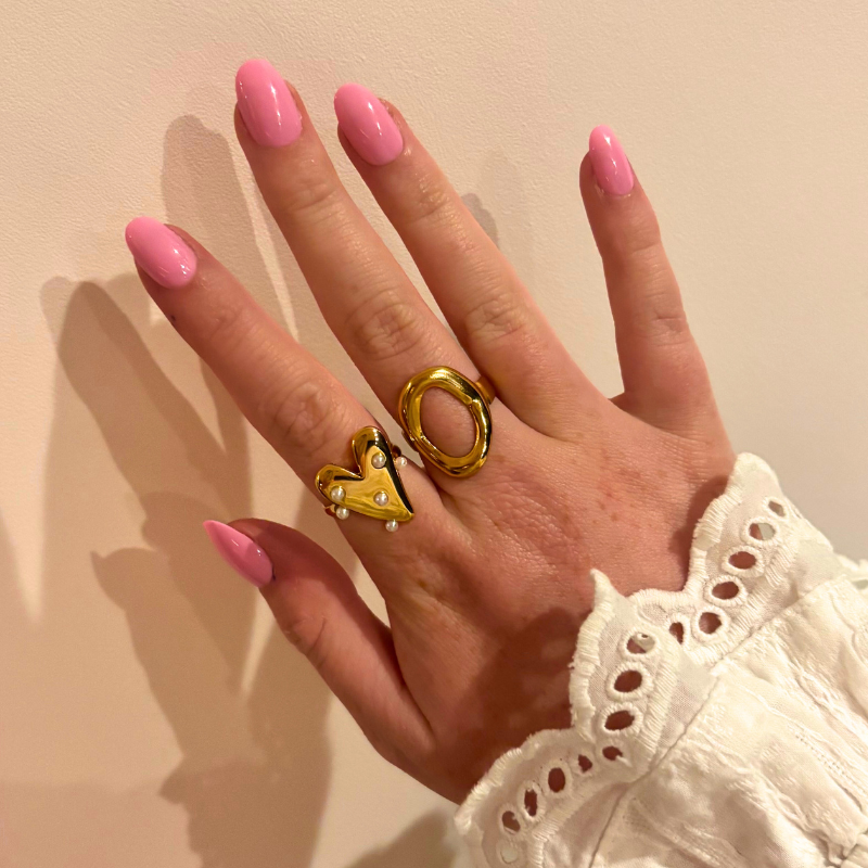 Hand met roze nagels die twee gouden ringen van FencyJewels draagt: hartvormige ring met parels en ovale statement ring