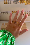 Hand met gouden ringen van Fencyjewels in hartvorm en golvend design, gedragen met groene blouse. Op de achtergrond diverse gouden en zilveren oorbellen op sieradenrek.