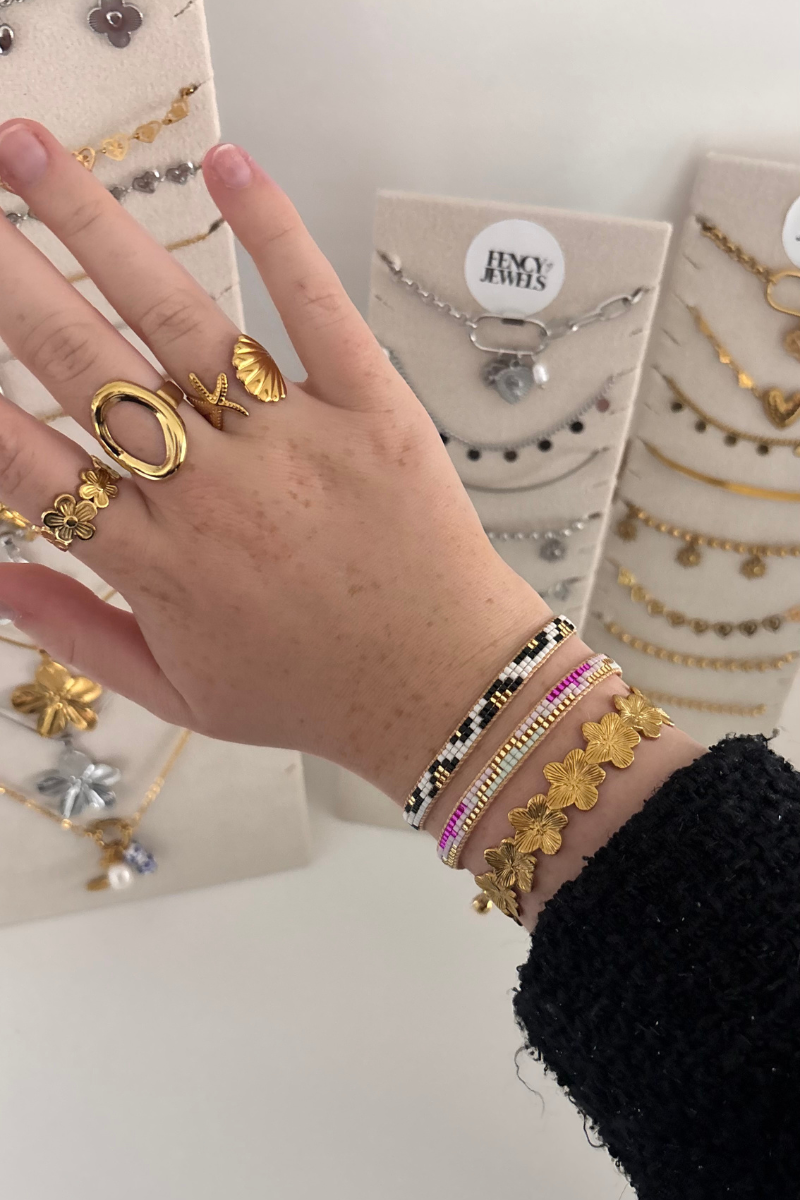 Hand met gouden ringen en armbanden van Fencyjewels, inclusief schelp- en bloemmotieven, gecombineerd met kleurrijke kralenarmbanden, trendy stainless steel sieraden voor jonge vrouwen.