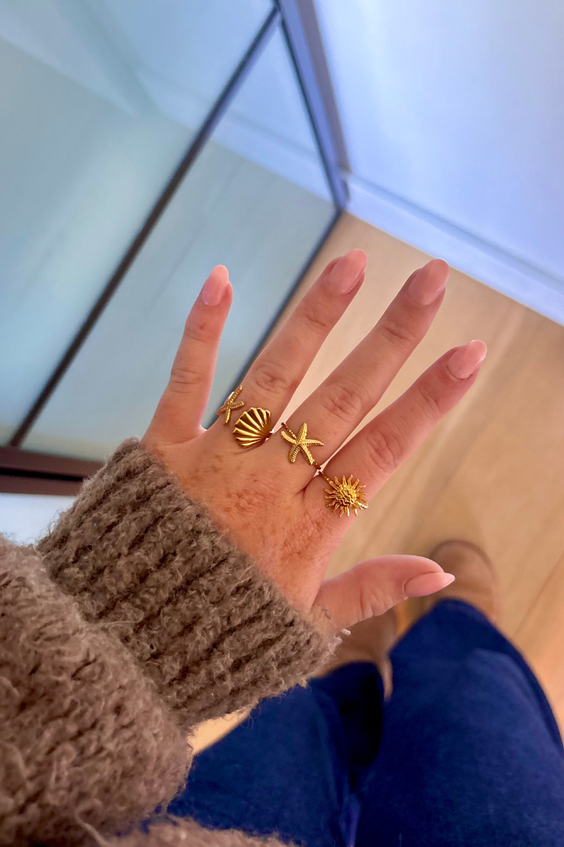 Hand met drie gouden ringen van Fencyjewels in schelp-, zeester- en zonontwerp, gedragen met bruine trui en jeans.