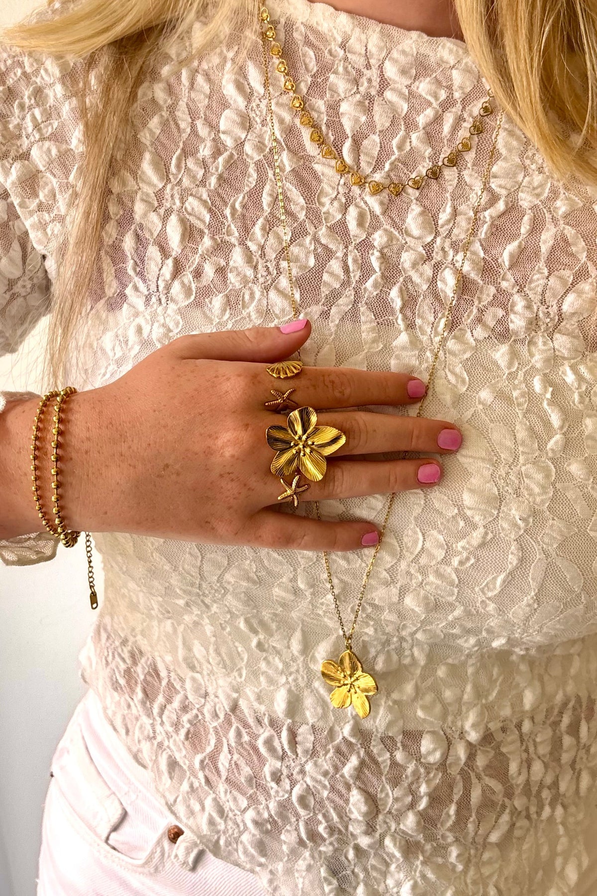 Gouden sieraden van Fencyjewels met bloem- en schelpringen en een bloemketting, gedragen op een witte kanten top met roze nagellak.